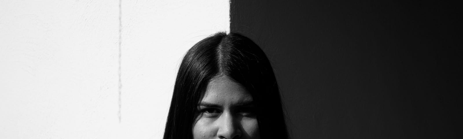 A black and white photo of a medium-skinned woman with long straight black hair parted in the middle. She is wearing a white, off-shoulder ruffled top with a dainty, heart-shaped necklace. She has a closed-mouth smile and is standing in front of a wall that is white on the left and black on the right, though it may be edited to look as such.