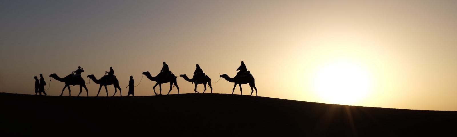 Three men on camels on horizon with two other camels in tow.