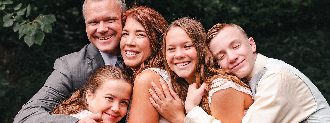 Wedding photo of a couple with three children around the ages of 9,11, and 14, all smiling and holding each other in a group embrace. They’re light-skinned, two femme-presenting kids with long reddish-brown hair, one masculine-presenting teen with short reddish-blonde hair, and the groom has short gray hair.