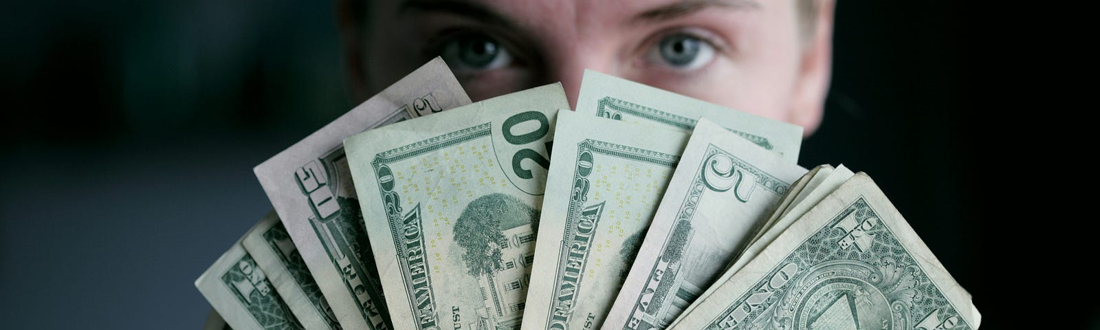 A woman holding several bank notes in a fan in front of her face. Her short nails are painted silver, and her eyes peek out from behind the money-fan.