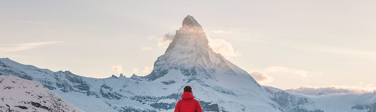 Person stands atop a mountain looking up at the sun rising from behind the Matterhorn mountain summit, in Europe.