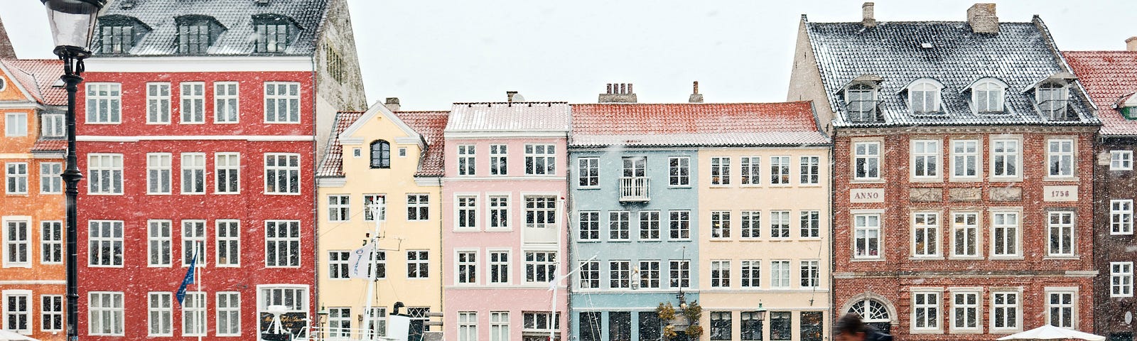 A scene from Copenhagen, showing some houses in Nyhavn from across the canal — a boat is in the canal and a bicyclist passing in front of the camera. It is winter and small amount of snow is coloring the roofs slightly white.