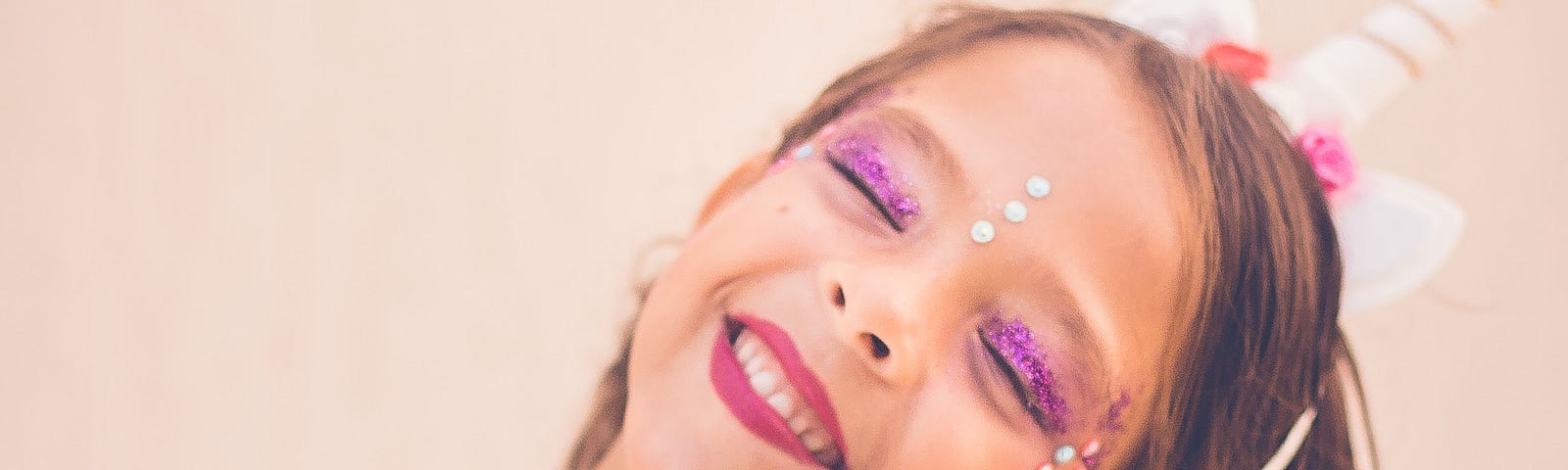A happy girl wears a unicorn horn headband, glittery purple eye shadow, and bright pink lipstick. She is also wearing face gems and a pink T-shirt with a unicorn motif.