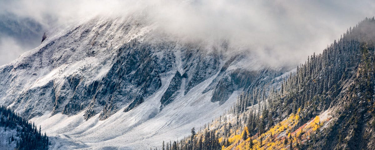 A rugged snow-dusted mountain arrayed in swirling clouds towers over a sweeping valley with evergreen trees and aspen changing from green to gold.