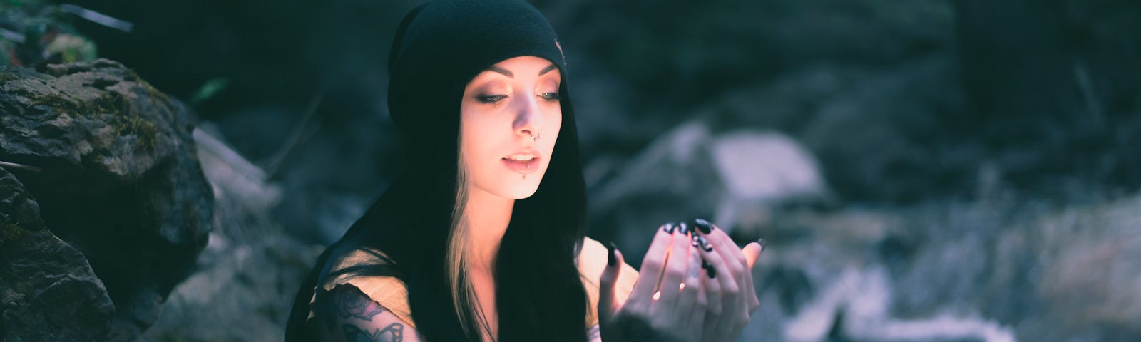 A woman cups her hands out in front of her chest. while she’s in fairly dark shade, her face and upper body are luminous. Mossy rocks and waterfall in the background.