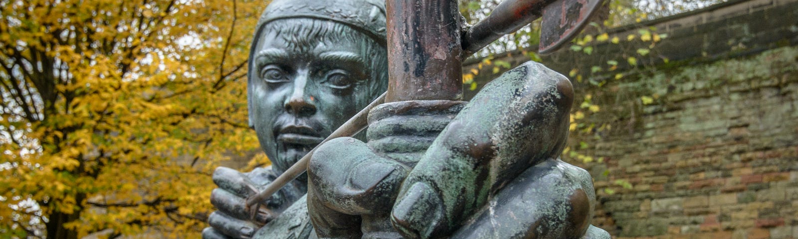 Weather-aged statue of an archer, his bow-hand in the extreme foreground as he aims his arrow with the other, ready to let it fly. Stone wall and trees with fall colors in background.