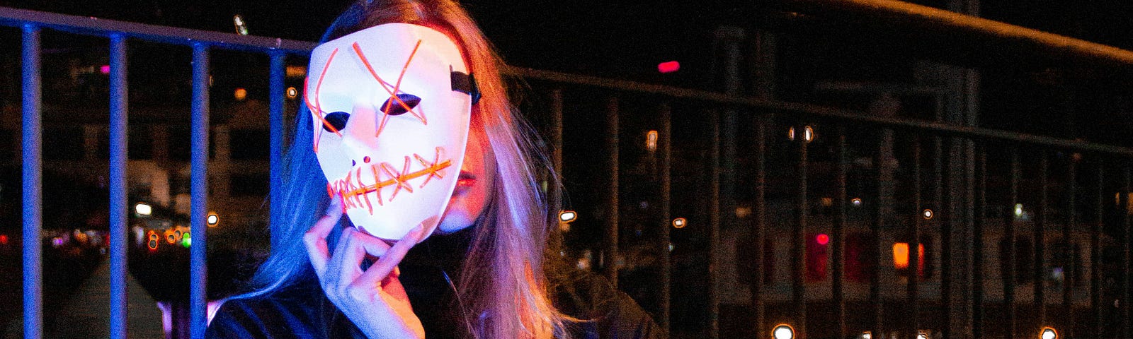 There’s a woman holding up a mask. It’s night and shes sitting on the pavement while leaning against a railing.