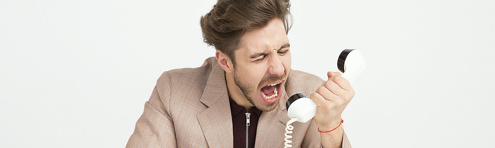 A white man yells angrily into a standing phone. He has sandy brown hair swooped back with a light amount of facial hair. He is wearing a tan blazer with a black quarter-zip sweatshirt underneath and an orange bracelet on his left wrist. On the white table he’s sitting at, there’s a pair of round frame black glasses sitting on the left and a white standing phone with black features on the right.