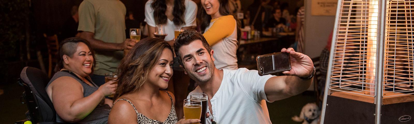 A group of friends in a restaurant take a selfie. They are holding drinks. Two of them are using wheelchairs.