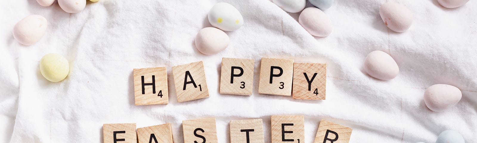 A flat surface with a white cloth on top decorated with pastel colored eggs and the words “Happy Easter” written out with Scrabble wooden tiles.