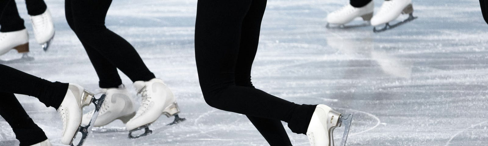 The legs and skates of a group of figure skaters wearing black leggings.