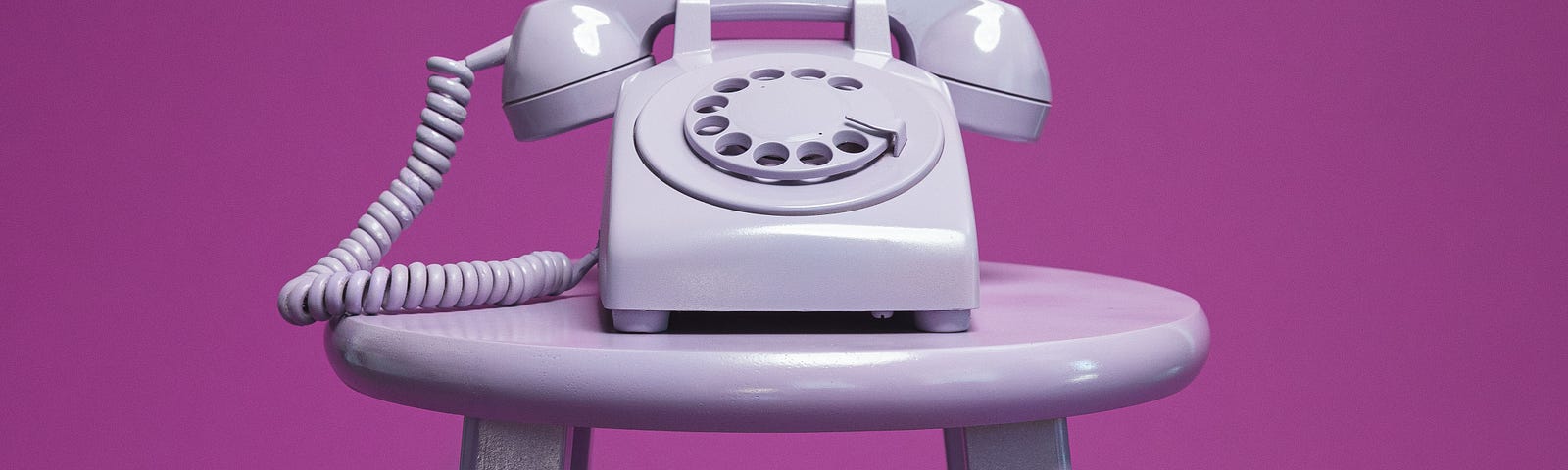 Old-style, white rotary phone atop white stool against purple background