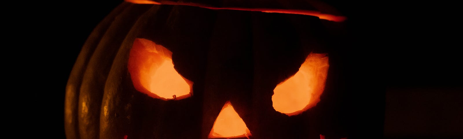 Image of a carved pumpking with mean eyes, and a mouth that lok like it’s been stiched, light shining from within.