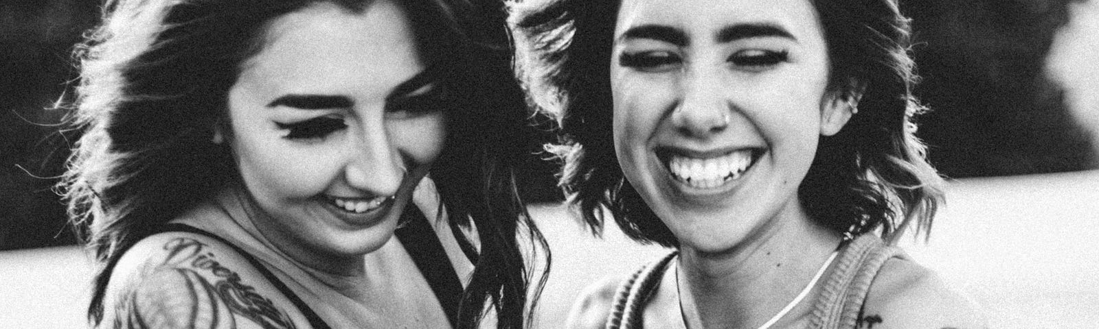 A laughing smiling couple of girlfriends in tatoos all in black and white, like a flashback.