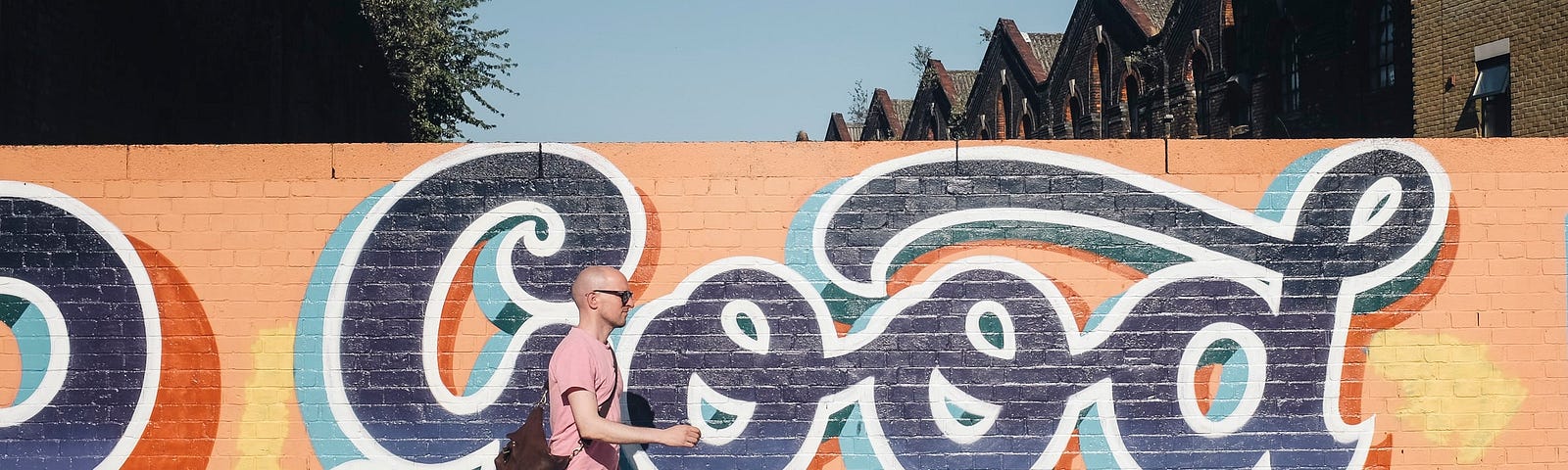 A man walks past a graffiti mural that says “Good” on a bright sunny day.