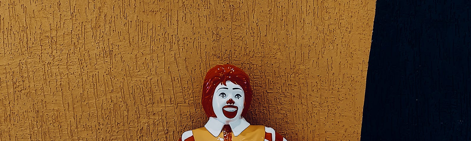 A statue of Ronald MacDonald sits on a bench, crossing his funny clown feet at the ankles.