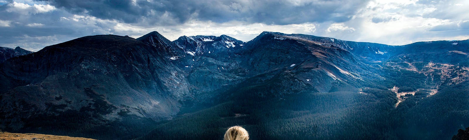 A woman sitting on a hilltop, deep in thought, gazing across an expanse of valley toward a mountain range