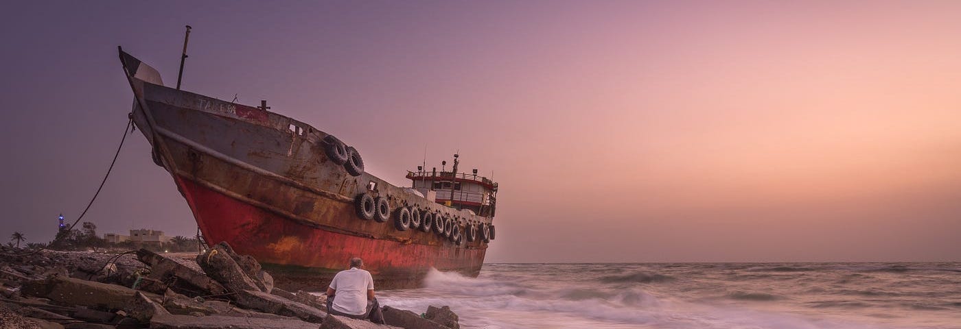 A man sits looking at a wrecked ship on the rocks
