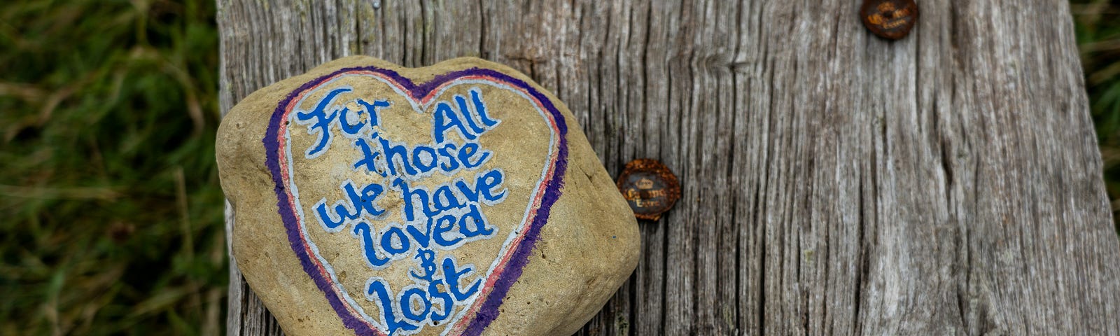 A large painted rock sits on a board. It has a pink and purple heart, the inside of which reads — For all those we have loved & lost — in blue curvy letters