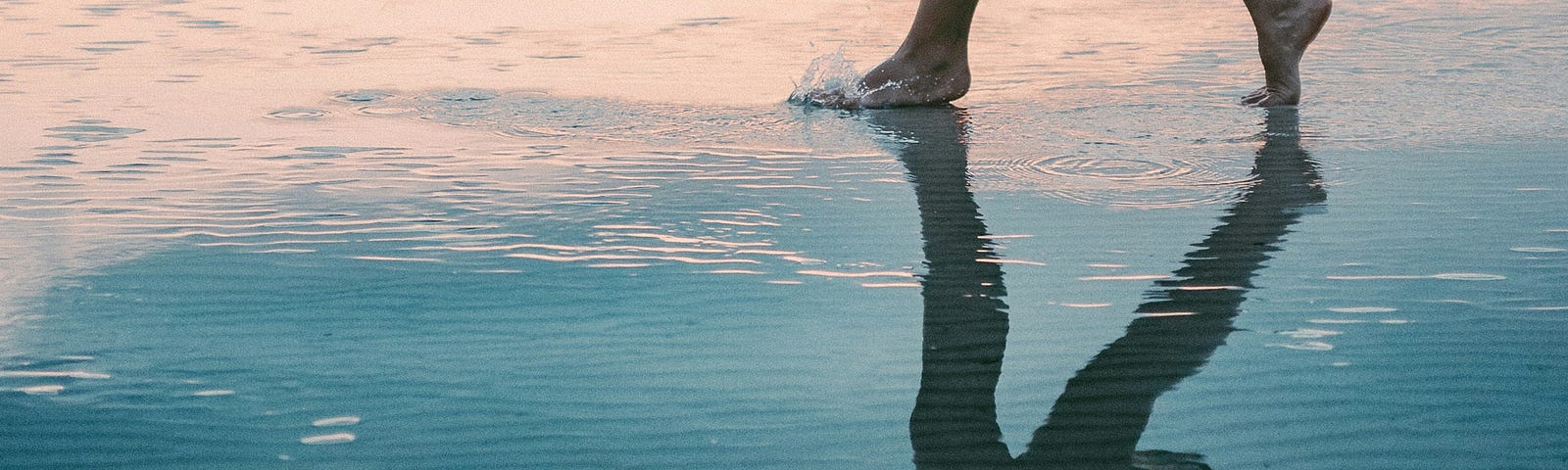 Photograph of a pair of women’s bare feet at the top of the image, walking on sand covered in blue water, with her feet, legs and outspread arms reflected in the water below.