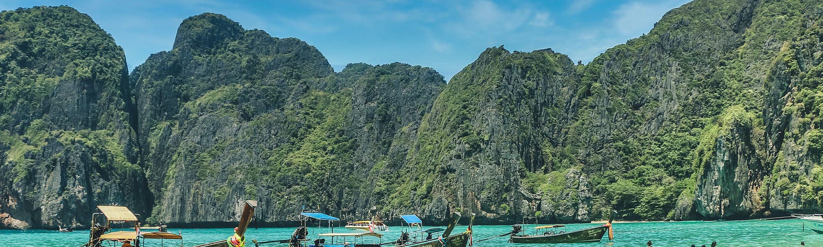 An image of a beach in Thailand, with traditional Thai longboats docked at the shore with mountains in the background.