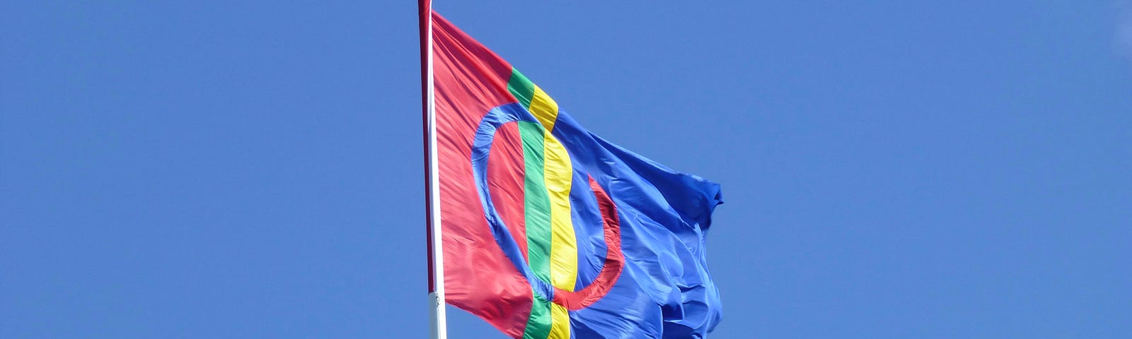 A Saami flag is pictured against a clear blue sky. The flag is red and blue halves separated by vertical sections of green and yellow. Superimposed over the background colors is a circle of blue and red.