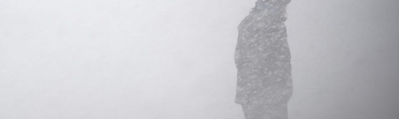 A man walking through a blizzard. You can only see his form through the snow.