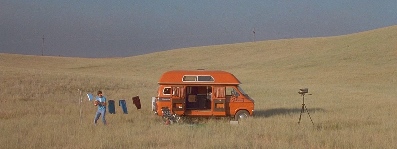 A man stands in a field next to the van he’s living in, holding a football, and recording himself with an old camcorder on a tripod.