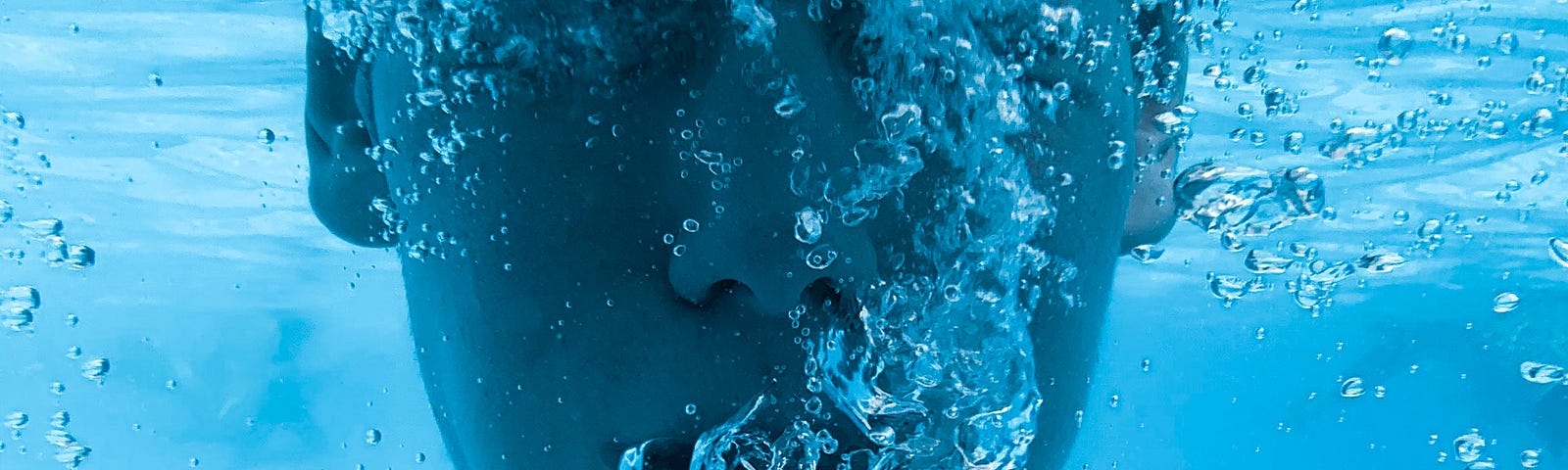 A young man is immersed in pool water. We see him from shoulders to eye level as he blows air bubbles out of his mouth.