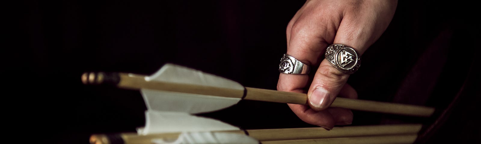 A hand wearing two signet rings grabs a wooden arrow from a quiver.