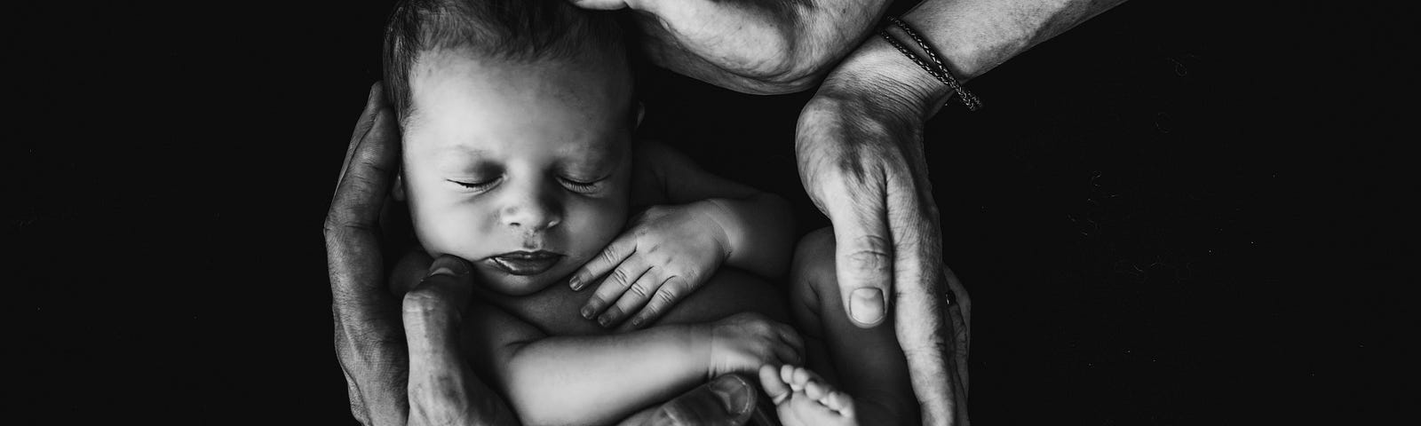 Mother’s Ode at Birth. Close up, black and white, of two sets of adult hands cradling a new infant.