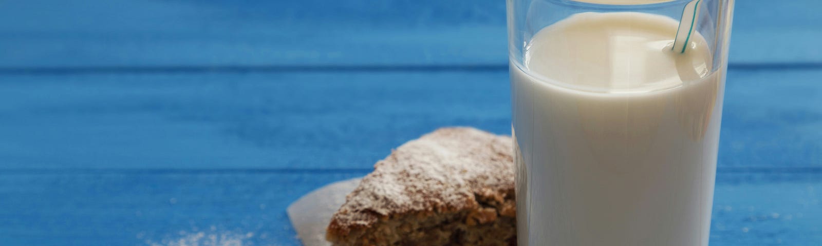 A glass of milk with a straw in it, placed on a blue wooden surface. Next to the glass is a piece of cake on a napkin, with some powdered sugar sprinkled around. The scene has a cozy and inviting feel