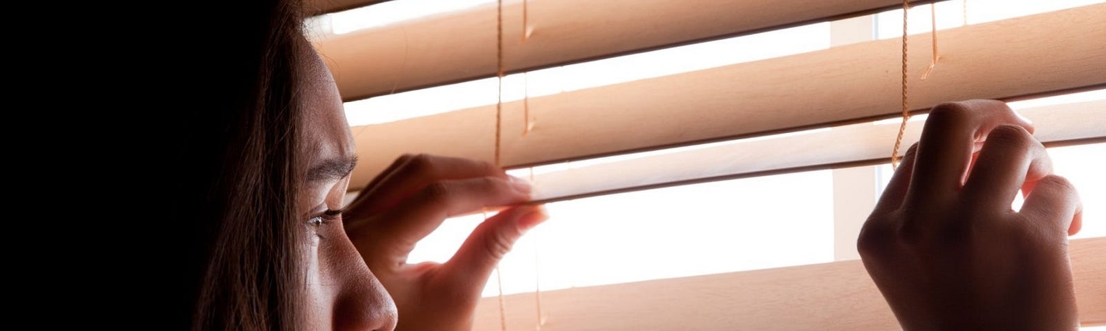 A woman looks out a window through blinds. Photo by lathuric/Getty Images