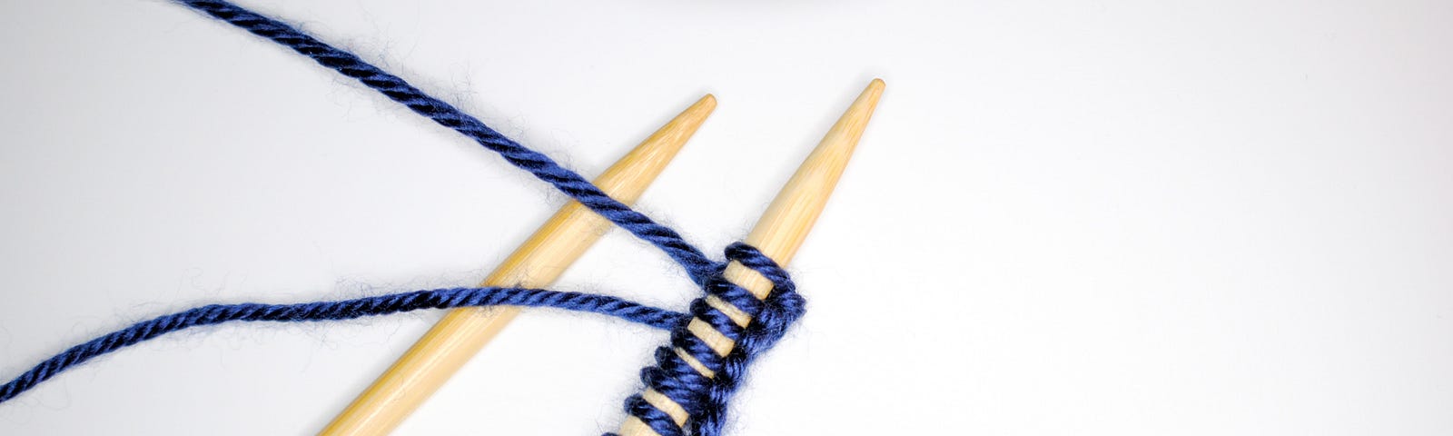 Two yellow knitting needles with several blue stitches cast onto them sit on a white background. A blue ball of wool is nearby.
