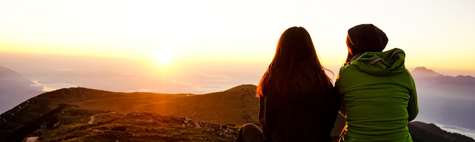 Two people sitting on a hillside watching a sunset in front of them.