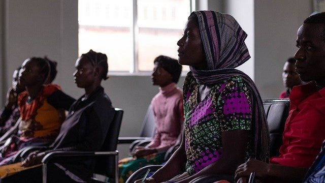 A group of black women in Tanzania sat in a classroom setting, looking attentively towards the front of the room. In the centre of the image is a woman in a pink patterned headscarf and a matching top