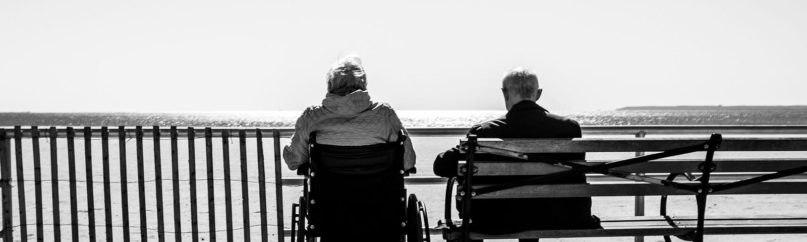 A couple sitting together, overlooking the water, one on a bench, the other in a wheelchair.