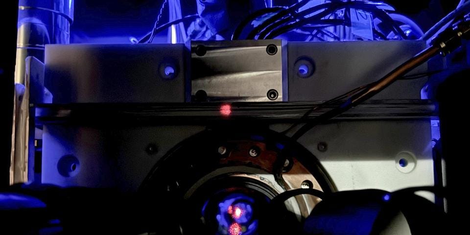 Close-up of a scientific instrument with wires and a circular metallic component, illuminated by blue light, featuring a small red dot at the center.