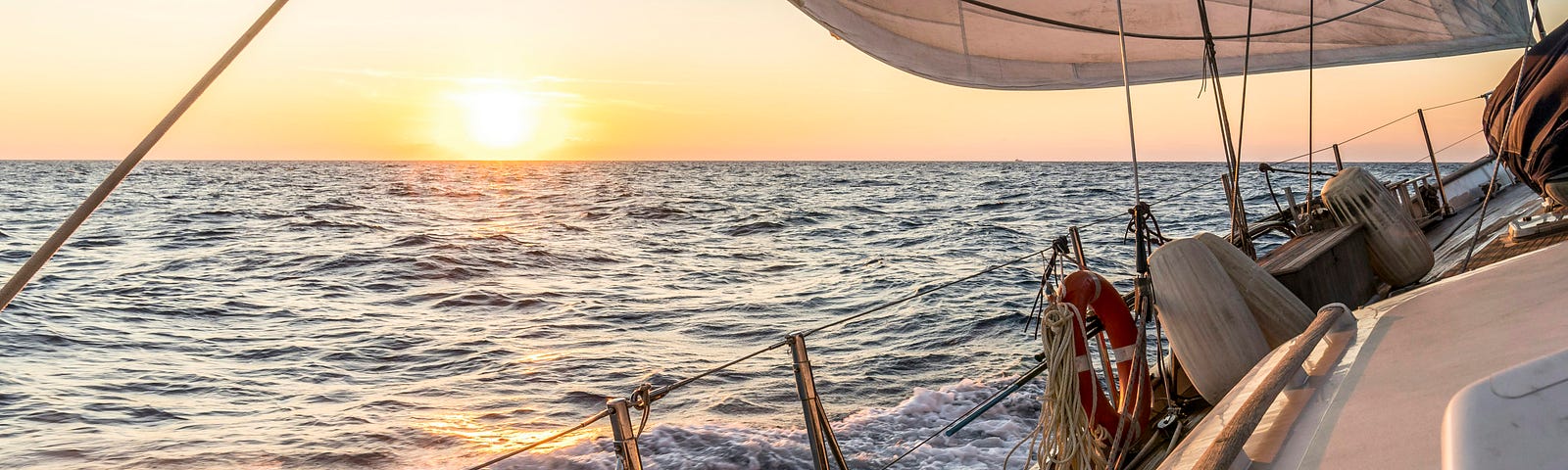 Part of a sailboat deck on the right, at sea with the sun setting on the horizon