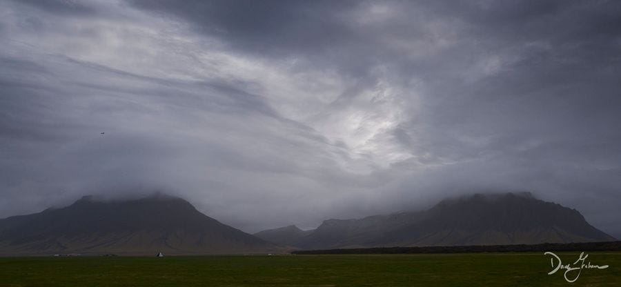 A cloudy sky over Icelandic mountains with green grass in the foregrounds and birds flying in between sky and earth.