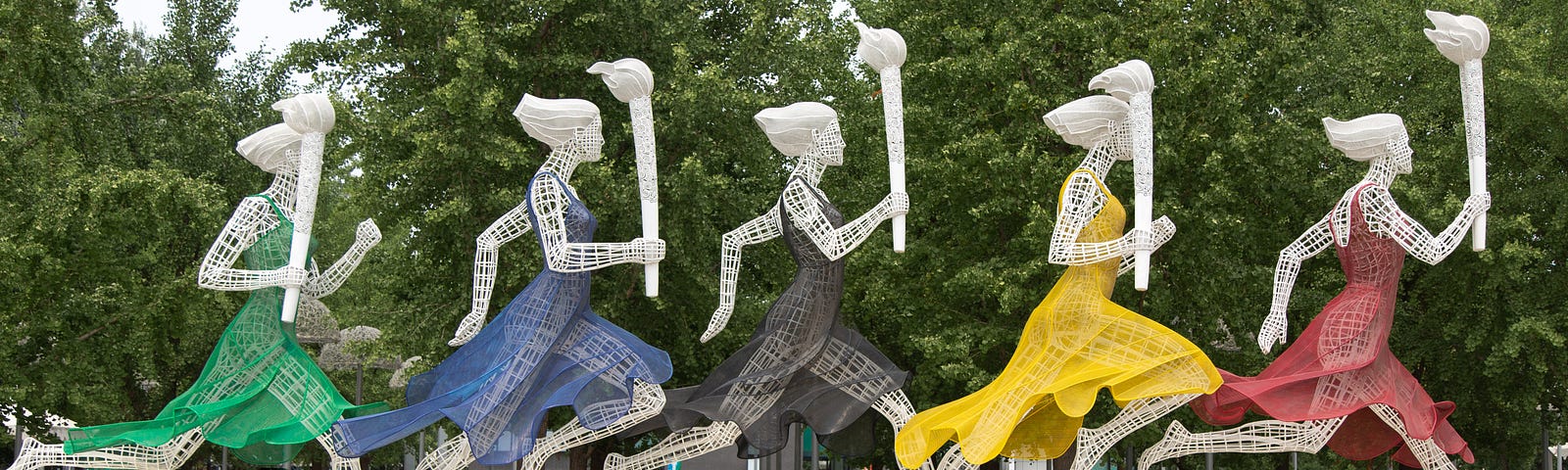 five wire sculptures of women dressed in green, blue, black, yellow, and red, each carrying the Olympic torch.