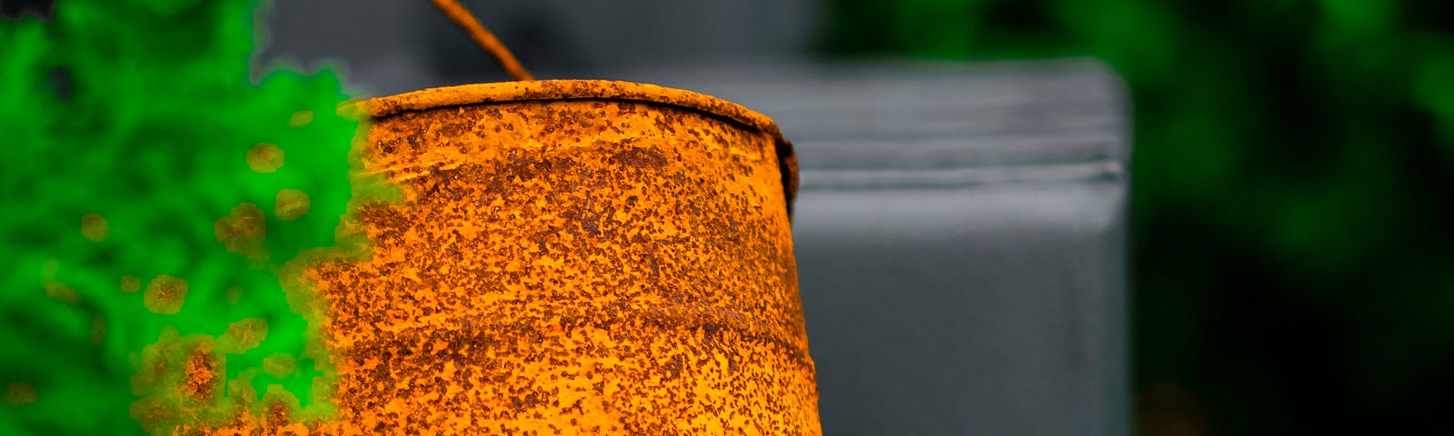 A rusty orange bucket hangs in front of a grey background behind a small sprig of bright green foliage.