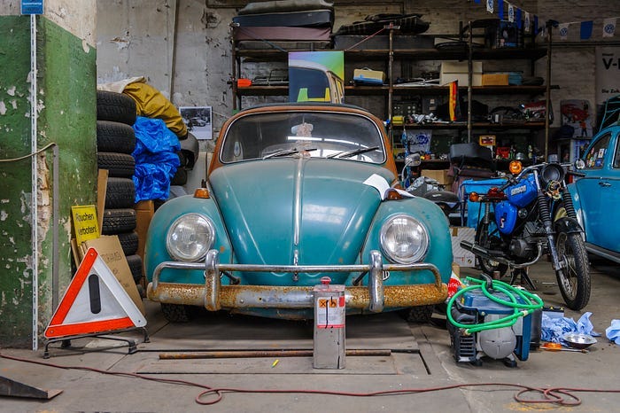 A rusty and dusty turquoise vintage VW Beetle sitting in a cluttered garage.