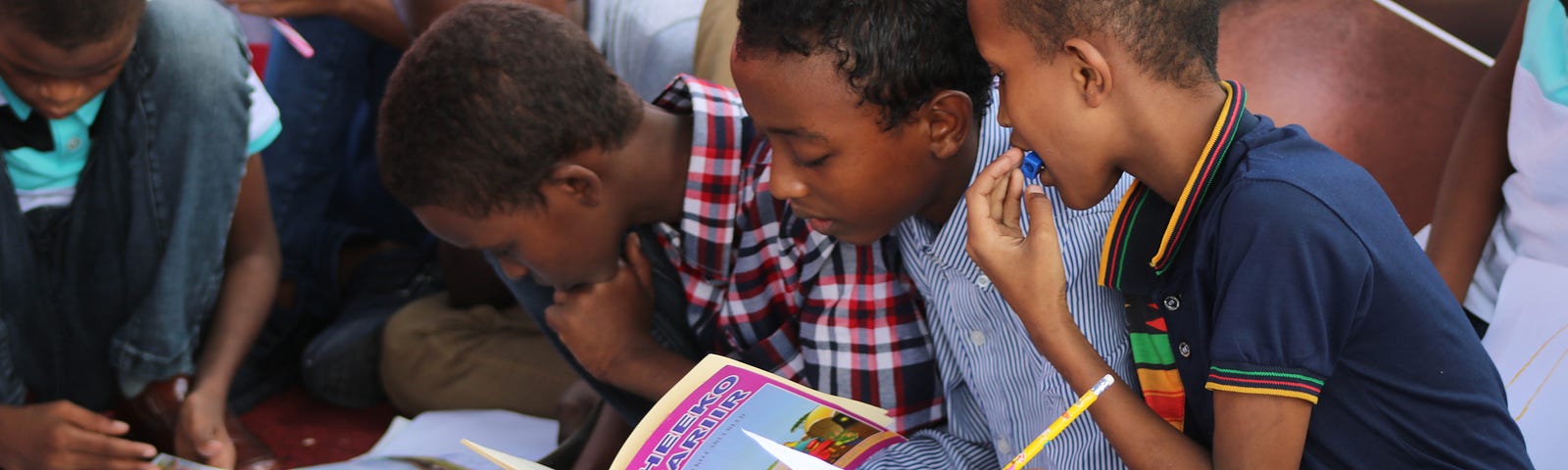 A group of children sit on the classroom floor learning, books and pencils in hand.