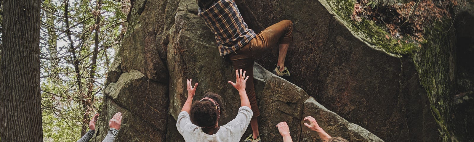 One person climbing the rock, and the other three stretch their arms and hands to support.