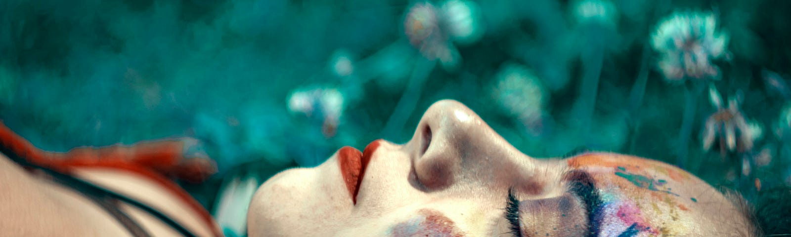 Young lady with red lipstick and coloured paint on one side lies in a field with eyes closed as if dreaming.