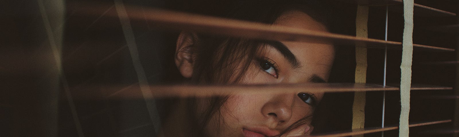 Girl with a bored expression on her face looks out through the brown blinds of a window as if longing for more.