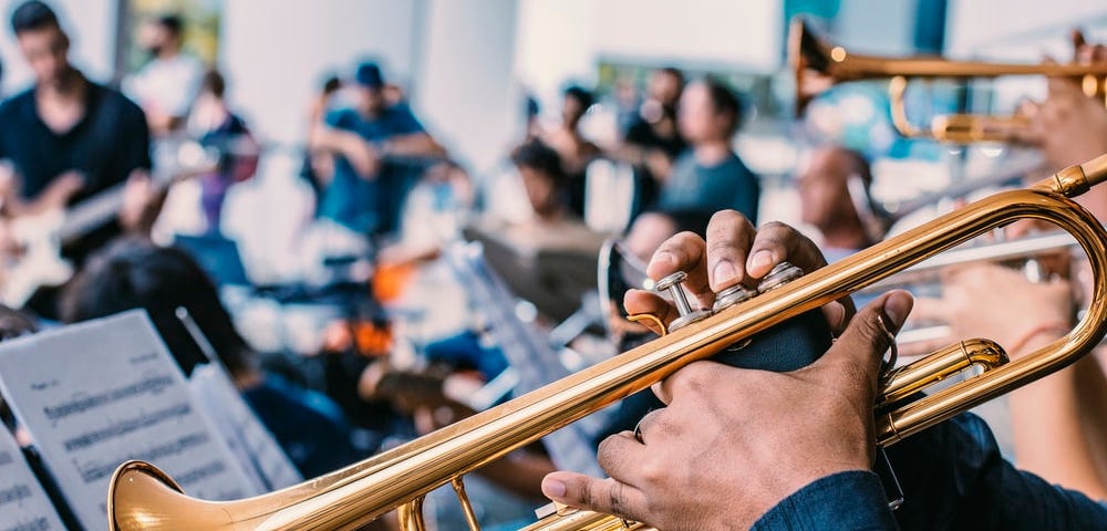 In the focus of the picture is a brass trumpet. A musician is playing music which can bee seen on a music stand and is pressing the first two valves. In the peripheral of the image are more musicians, including more trumpet players, percussionists, and a guitar player.