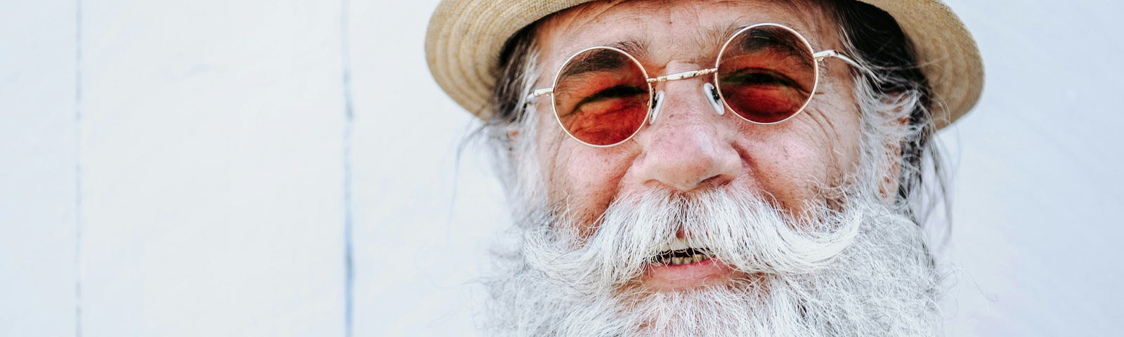 A smiling older man with a straw hat, round sunglasses, meticulously teased-up white mustache and a bushy white beard. He has no shirt on, just some decorative beads. He looks happy and relaxed, like Santa on vaccation.