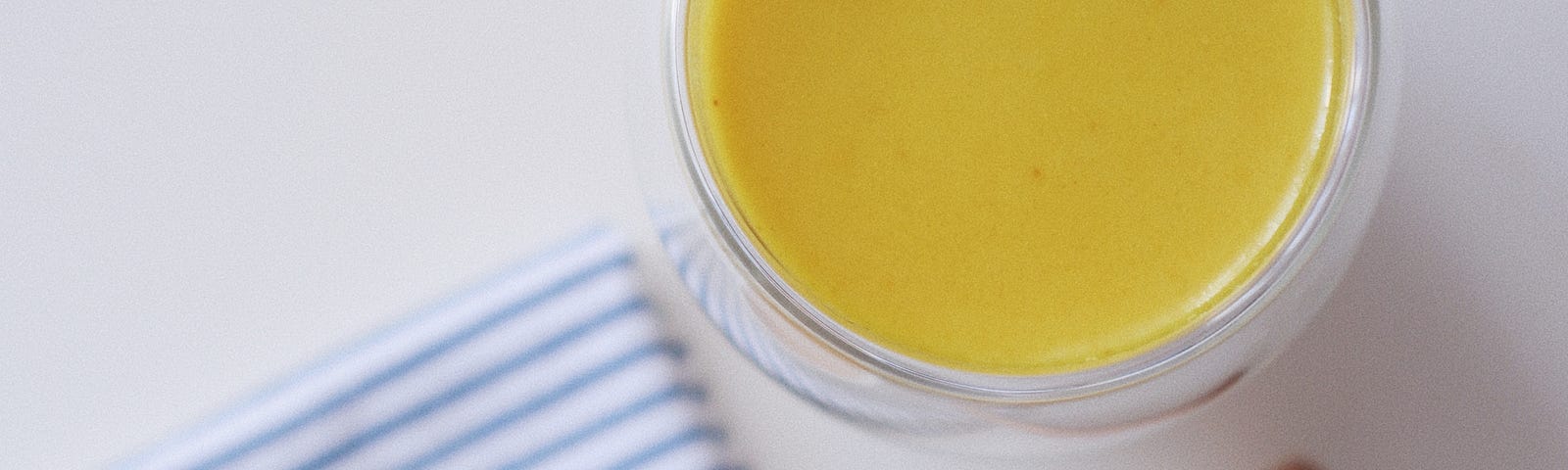 There is a heart-shaped small white dish containing an orange liquid at the lower left. It sits on a blue and white striped cloth. At the top right there is a cup of a mustard-colored liquid.